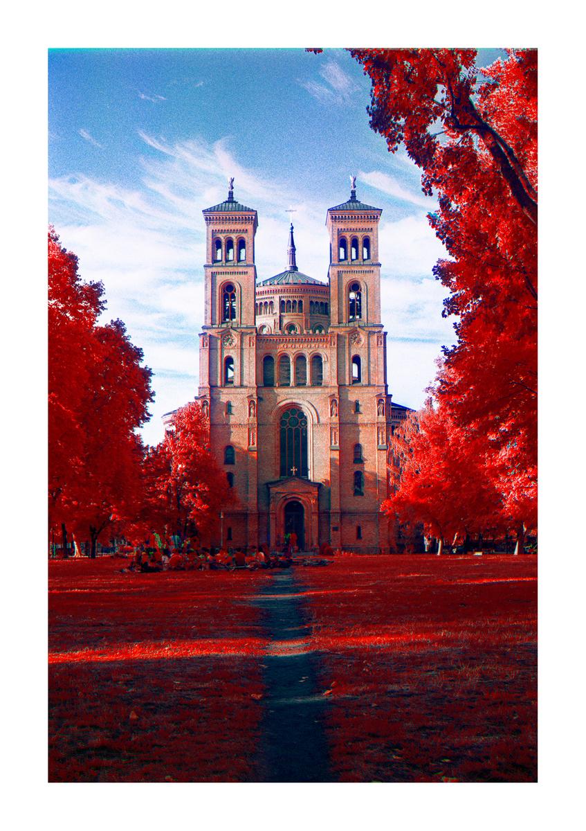 A path leads through a red field to a large church with two towers under a blue sky.