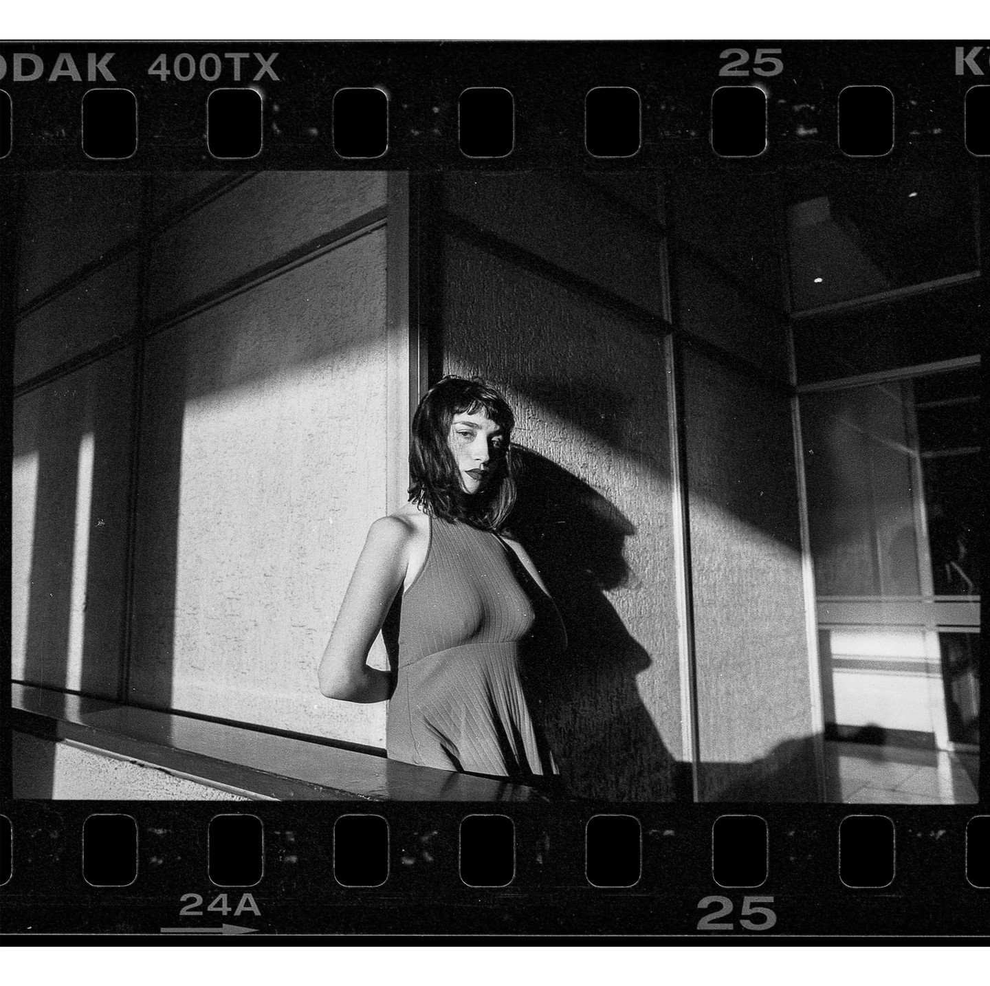 A woman with dark hair and dark lipstick stands in a sleeveless dress against a building wall, with strong shadows cast behind her.