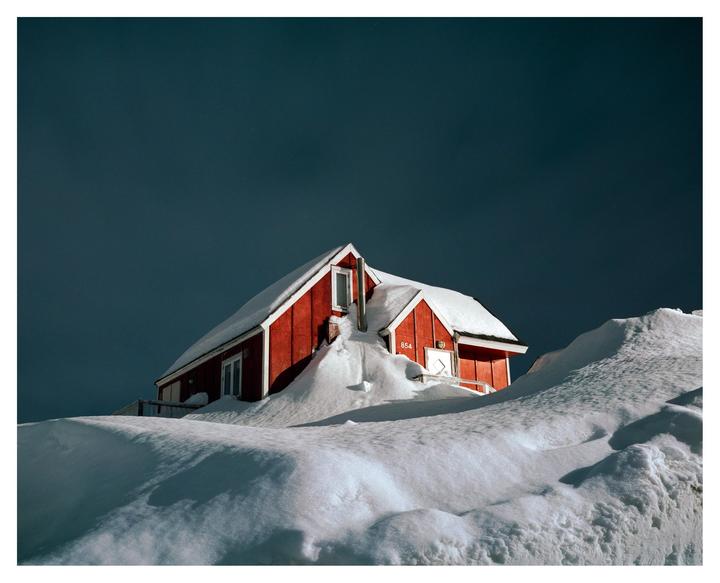 A red house with a snow-covered roof is nestled in deep snowdrifts under a dark sky.