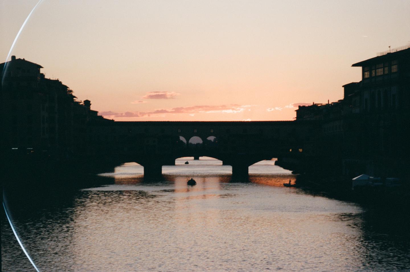 A river flows under a bridge with buildings on either side, silhouetted against a colorful sunset sky.