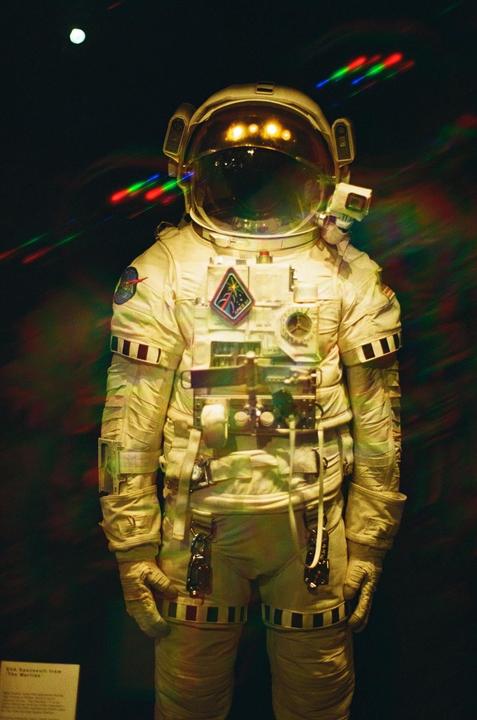 An astronaut suit stands in a dark room with colorful light streaks and a bright orb in the background.