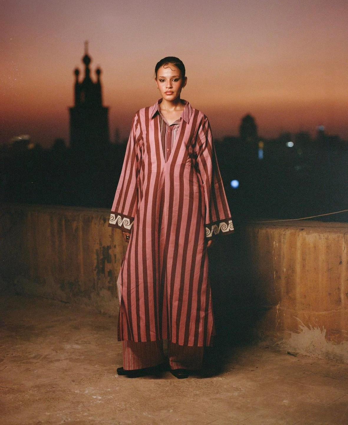 A woman in a striped tunic and pants stands on a rooftop at dusk with a city skyline in the background.