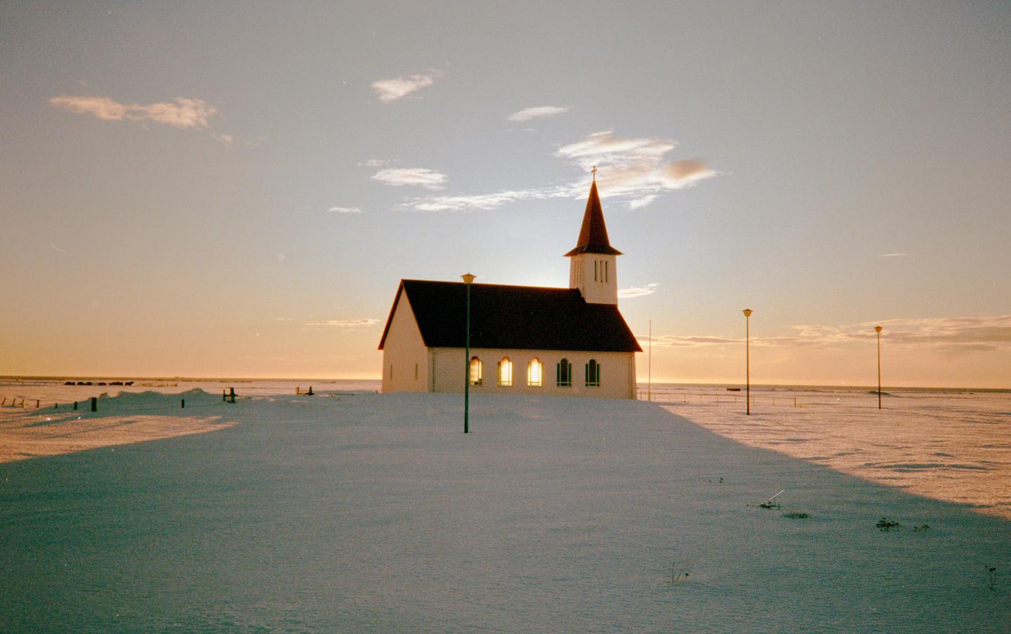 A white church with a red roof and steeple stands in a snow-covered landscape under a golden hour sky.