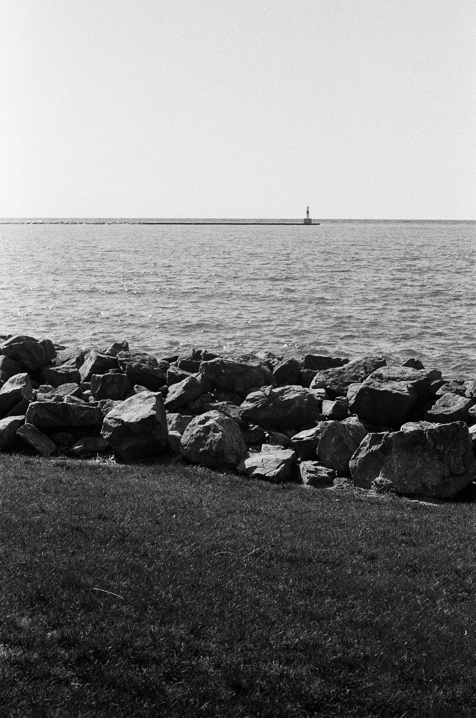 A lighthouse stands on a distant pier across a body of water, with rocks and grass in the foreground.
