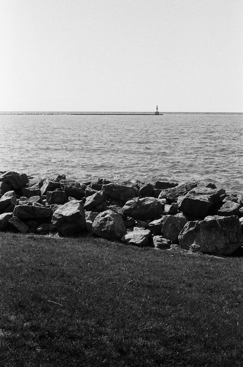 A lighthouse stands on a distant pier across a body of water, with rocks and grass in the foreground.