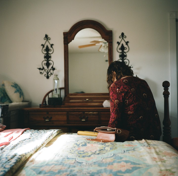A woman sits on a bed in front of a wooden dresser with a mirror.