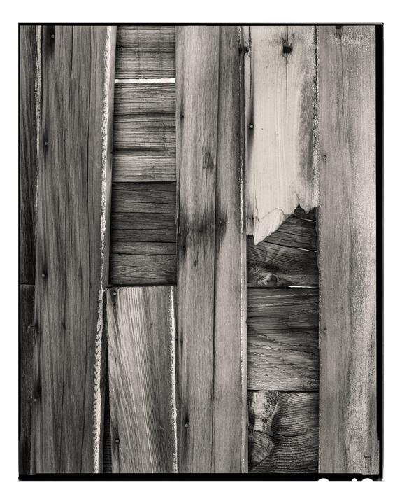 A black and white close-up shows a weathered wooden fence with several planks missing or broken, revealing more wood behind.