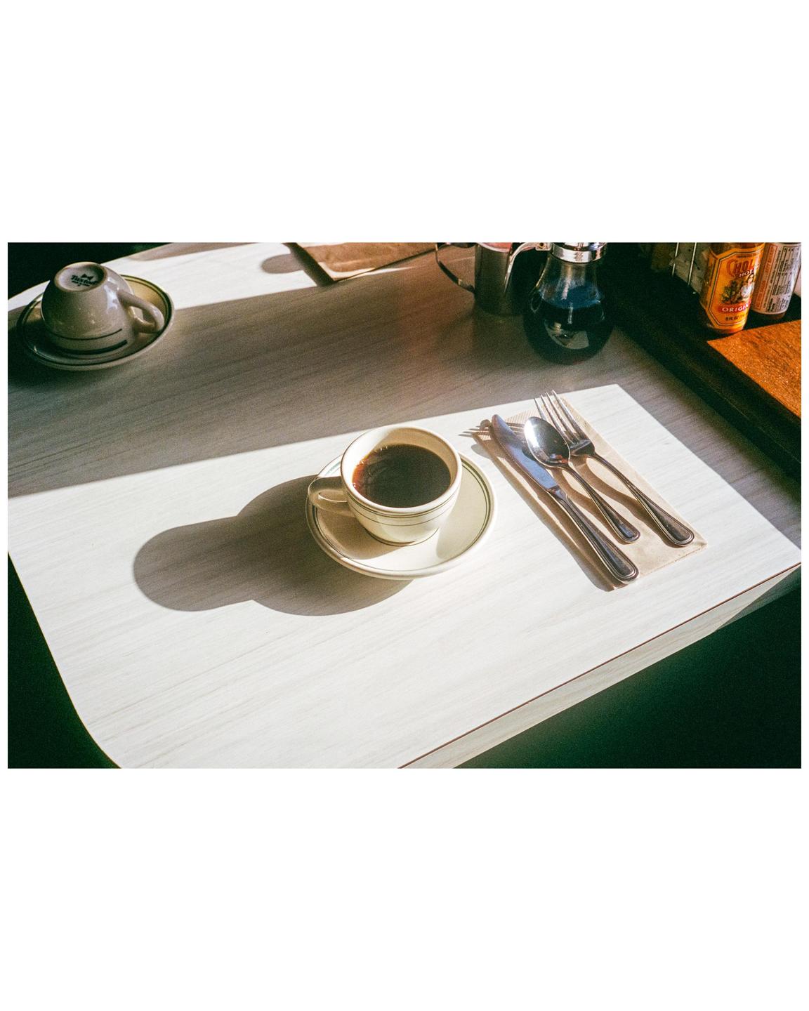 A cup of coffee and cutlery are arranged on a table, illuminated by a strong diagonal beam of sunlight.