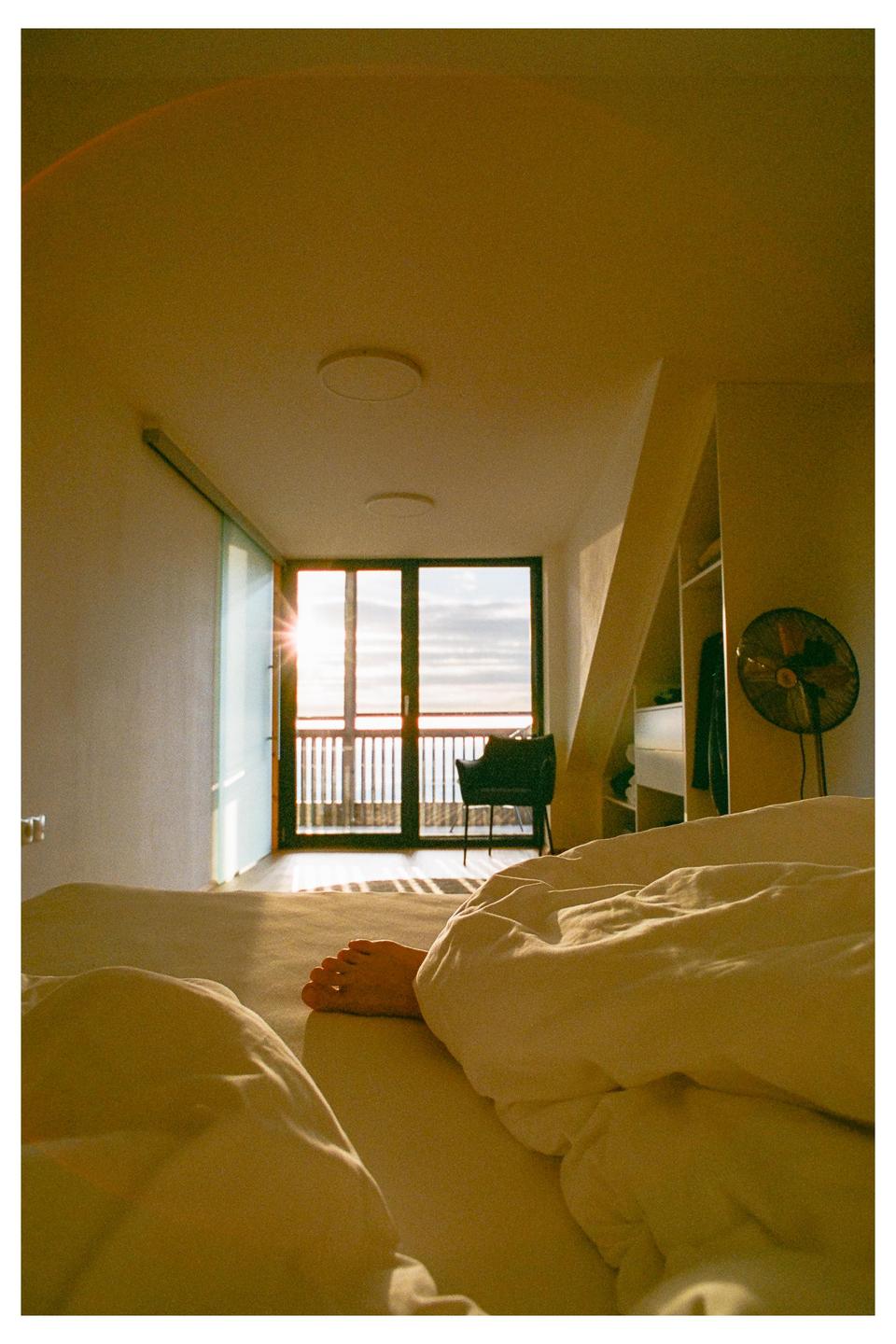 A foot is visible on a bed in a sunlit room with a balcony overlooking the ocean.