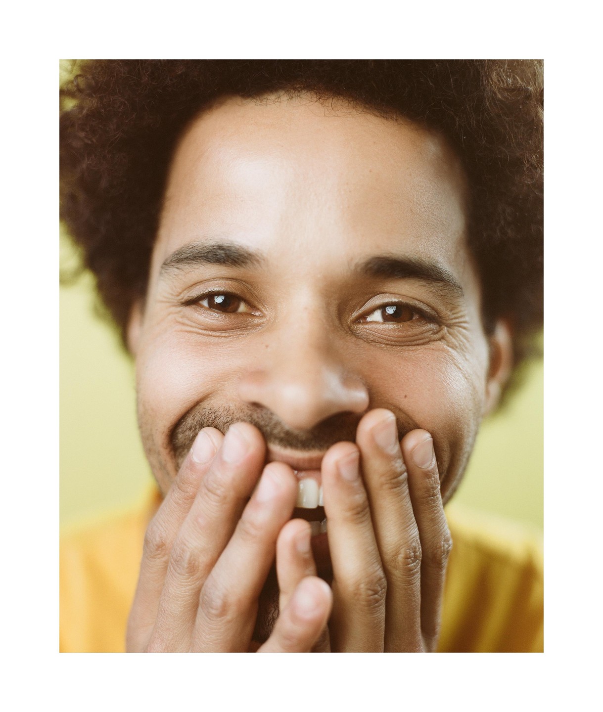 A man with curly hair smiles with his hands covering his mouth.