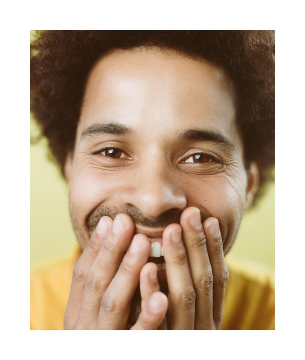 A man with curly hair smiles with his hands covering his mouth.