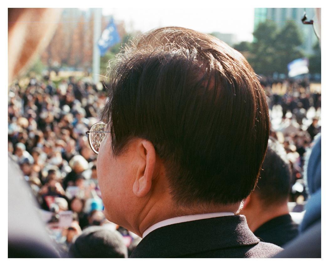 A man with glasses stands in a crowd of people, looking away from the camera.