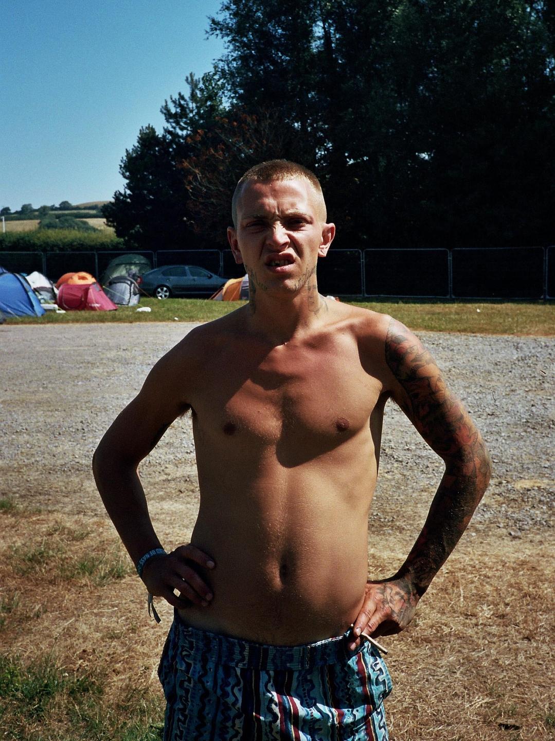 A man with a shaved head and tattoos stands shirtless with his hands on his hips, making a face at the camera.