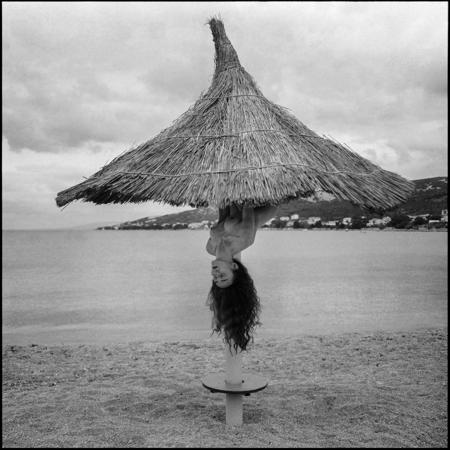 A woman hangs upside down from a straw umbrella on a beach next to the sea.