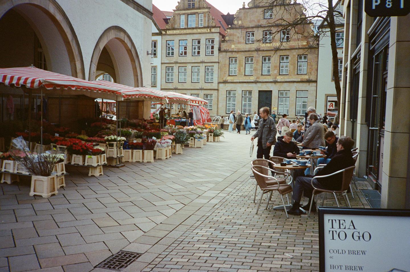 A flower market with red and white striped awnings is set up on a cobblestone street next to a cafe with people sitting at tables.
