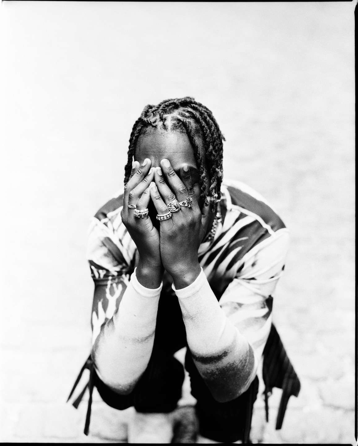 A man with braided hair covers his face with his hands, which are adorned with multiple rings.