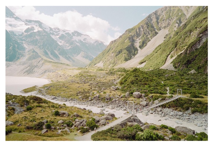 A suspension bridge crosses a river in a valley with mountains and a lake.