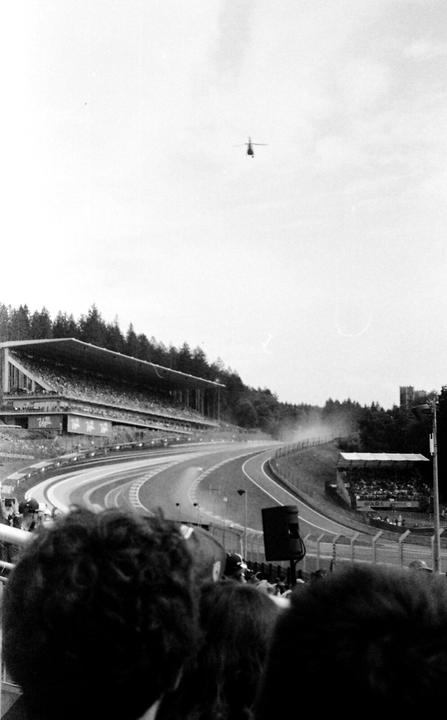 A helicopter flies over a race track with spectators in the stands and trees in the background.