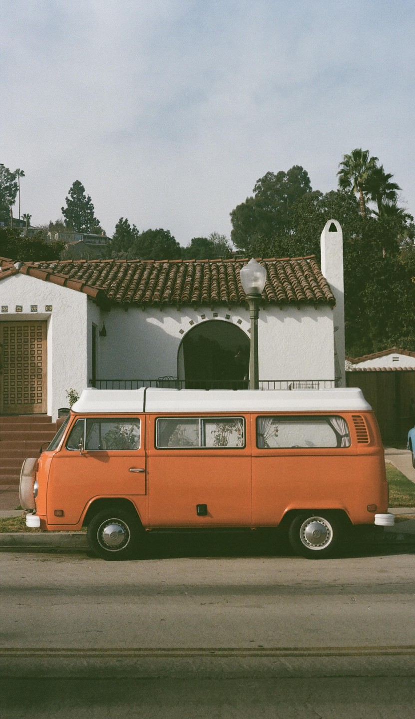 An orange van is parked on a street in front of a white house with a tiled roof.