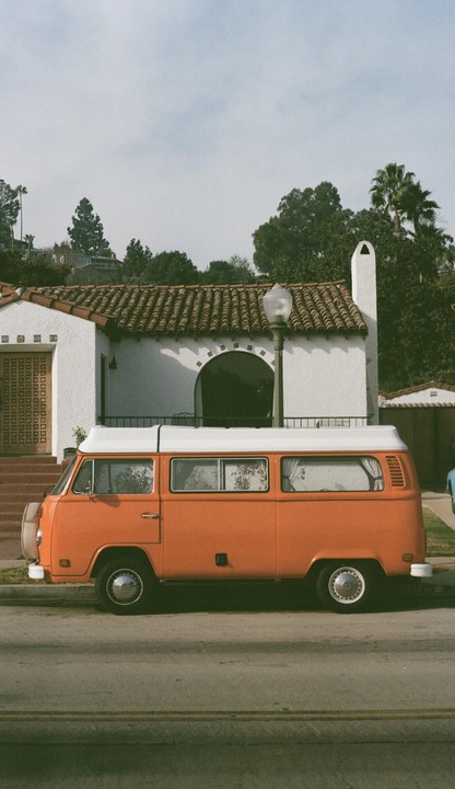 An orange van is parked on a street in front of a white house with a tiled roof.