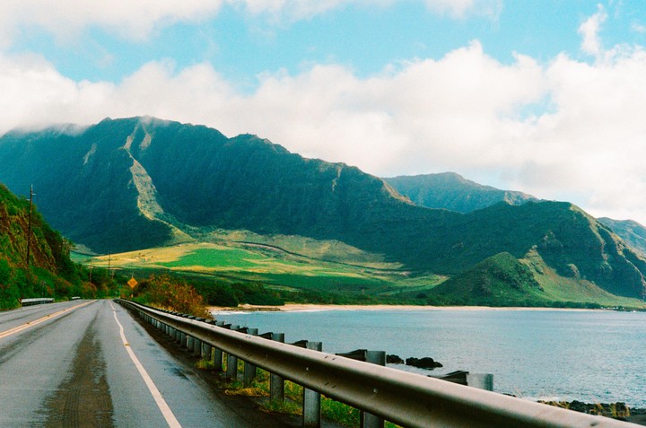 A coastal road runs alongside the ocean with lush green mountains in the background under a partly cloudy sky.