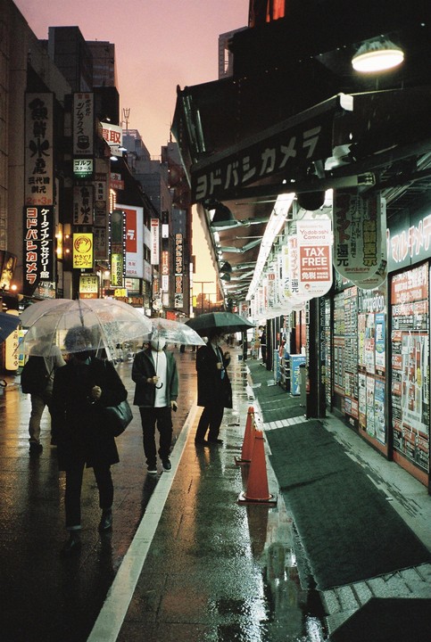 People walk with umbrellas on a wet street lined with neon signs and storefronts in Shinjuku, Tokyo.