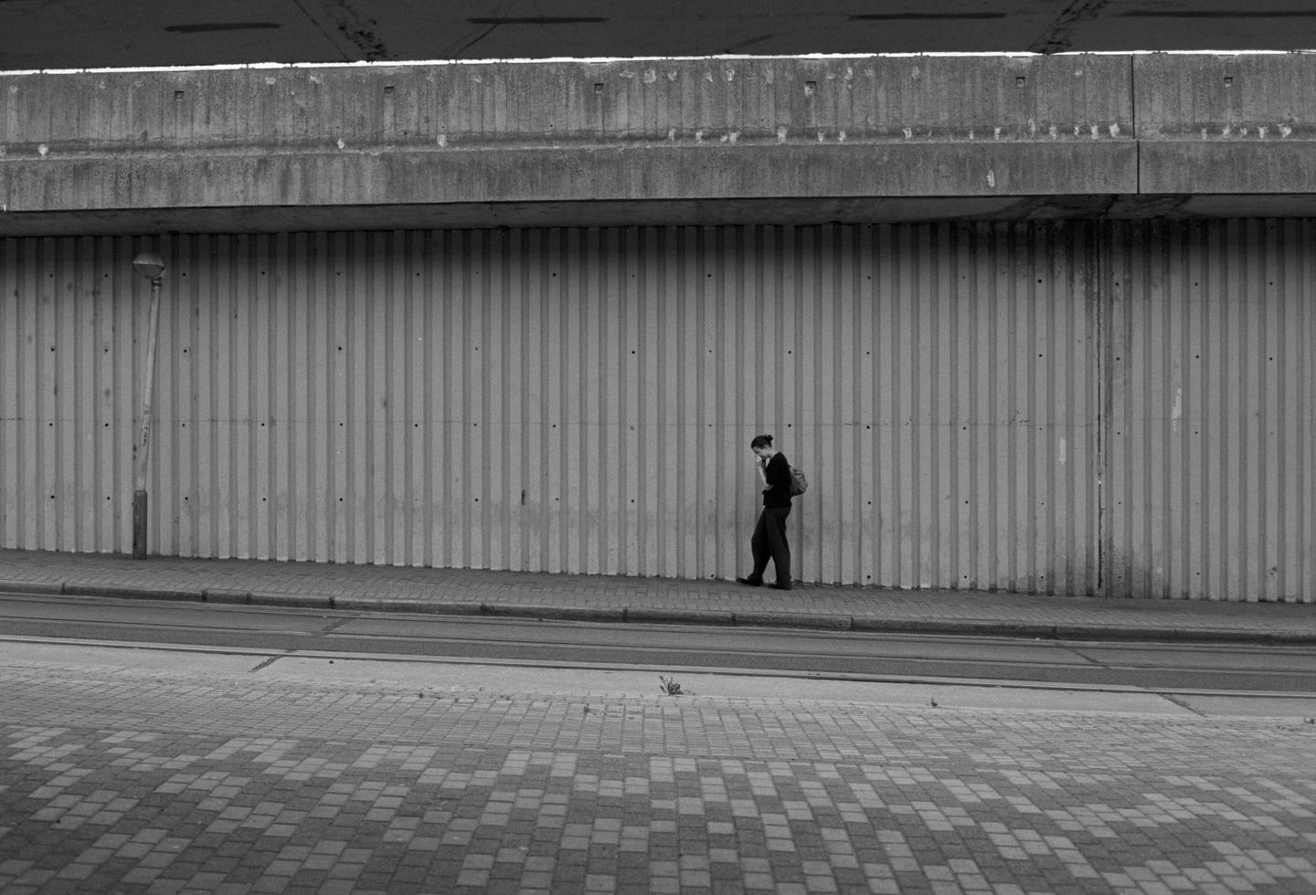 A woman walks along a sidewalk next to a corrugated wall under a concrete overpass.