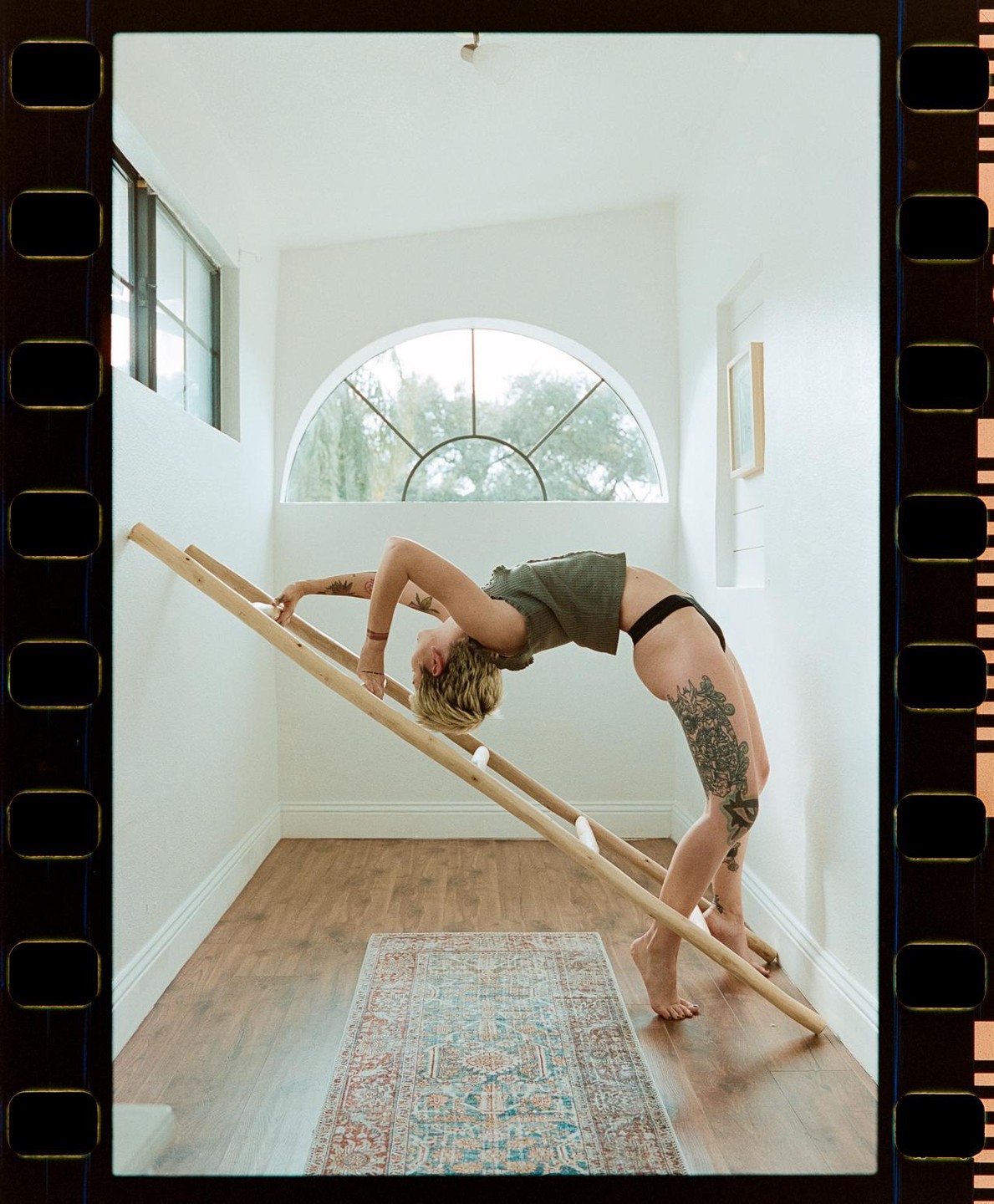 A woman with tattoos on her leg and arm bends backward over a wooden ladder in a room with a patterned rug on the floor.