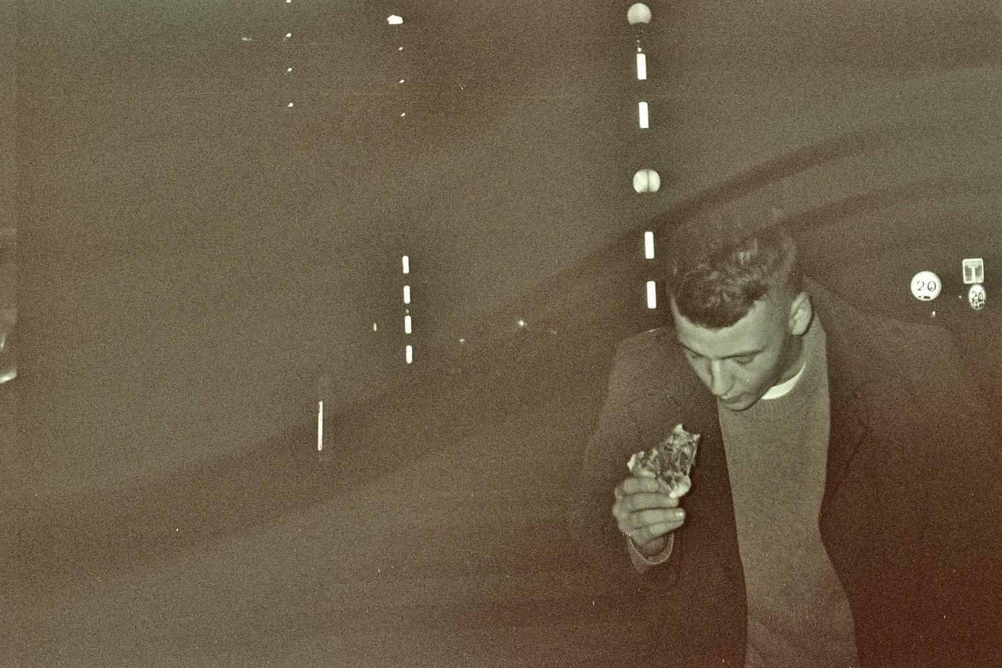 A man in a coat and sweater holds a piece of pizza at night.