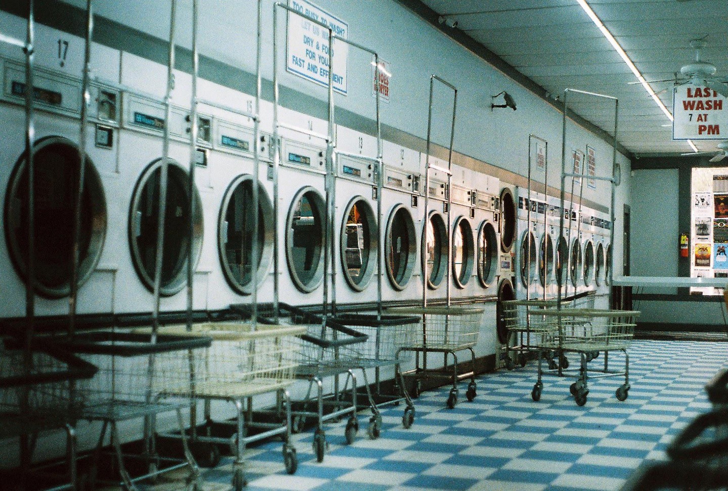 A row of washing machines and laundry carts are visible in a laundromat with a blue and white checkered floor.