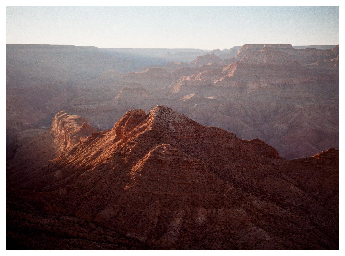 A vast canyon landscape is illuminated by the setting sun, casting long shadows across the rugged terrain.