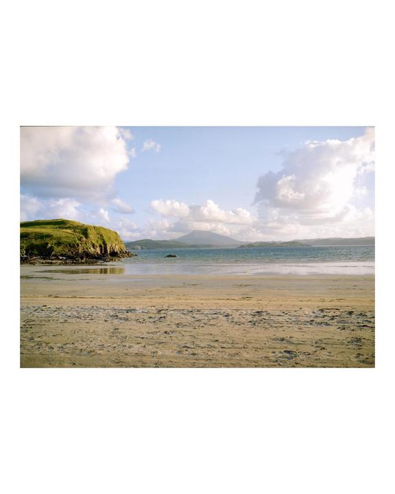 A sandy beach with a grassy hill on the left and a mountain in the distance under a cloudy sky.