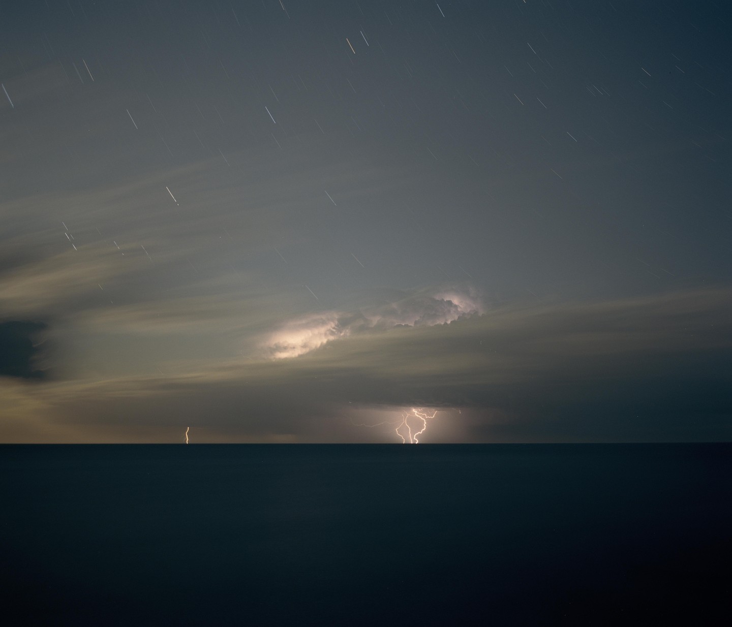 Lightning strikes over the ocean under a cloudy night sky with star trails.