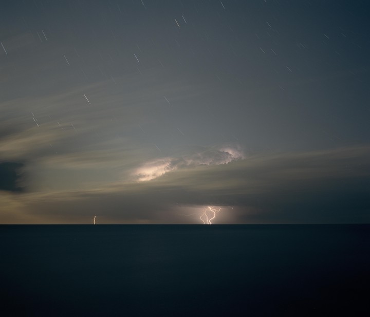 Lightning strikes over the ocean under a cloudy night sky with star trails.