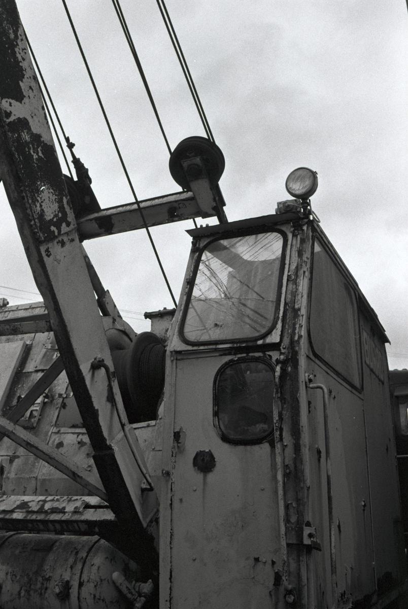 A black and white photograph shows the weathered cabin and boom of a large piece of industrial machinery against a cloudy sky.
