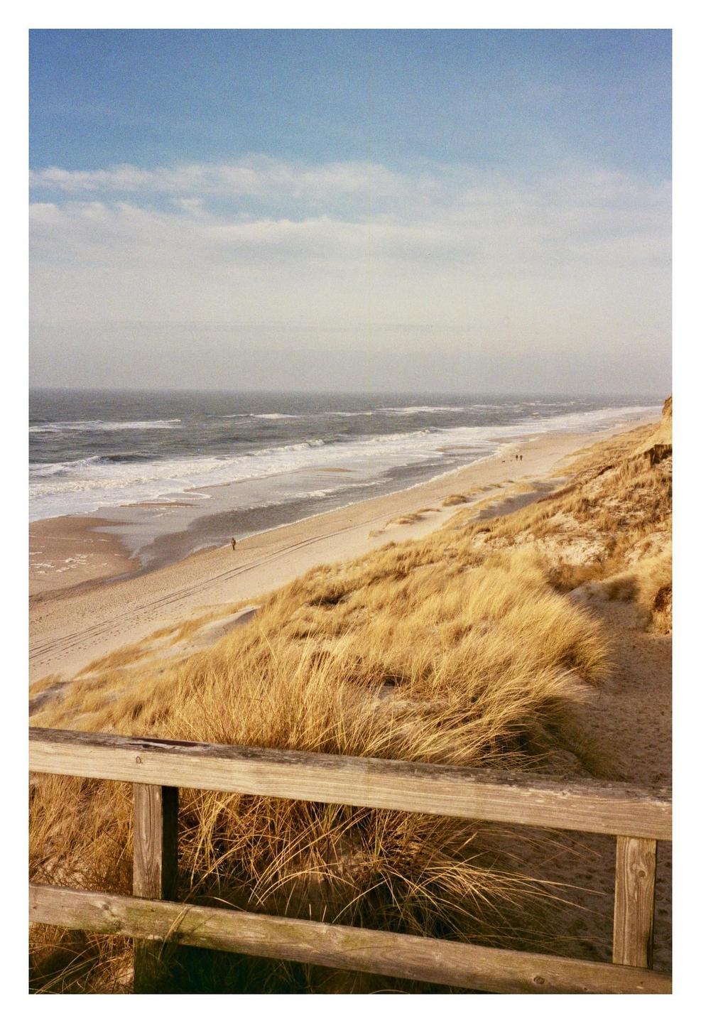 A wooden fence overlooks a sandy beach with dry grass, ocean waves, and a hazy sky.