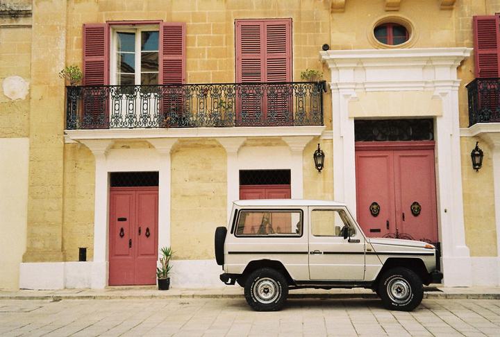 A white SUV is parked in front of a building with pink doors and shutters.