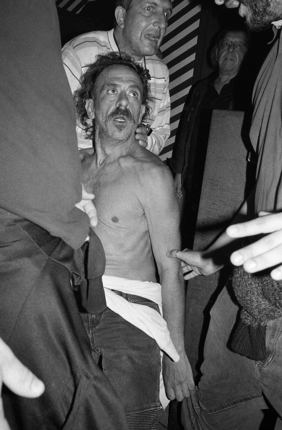 A shirtless man with a mustache is surrounded by other men, one of whom has his hands on the shirtless man's shoulders.