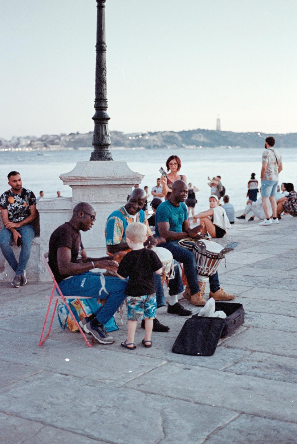 Three men play drums for a child on a stone street next to a body of water in Lisbon.