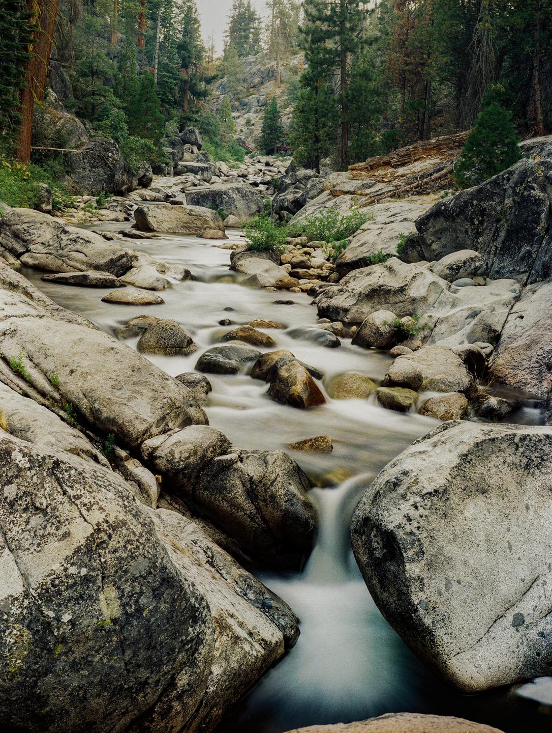 A long exposure shot shows a river flowing over and around large rocks in a forest.