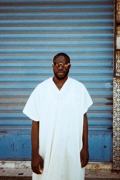 A man wearing a white tunic and sunglasses stands in front of a blue corrugated metal door next to a patterned tile column.