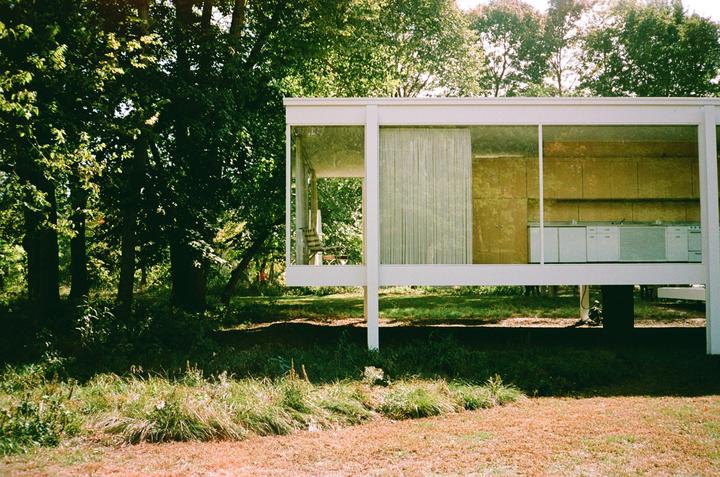 A modern glass house with a kitchen visible through its windows stands among green trees and grass.