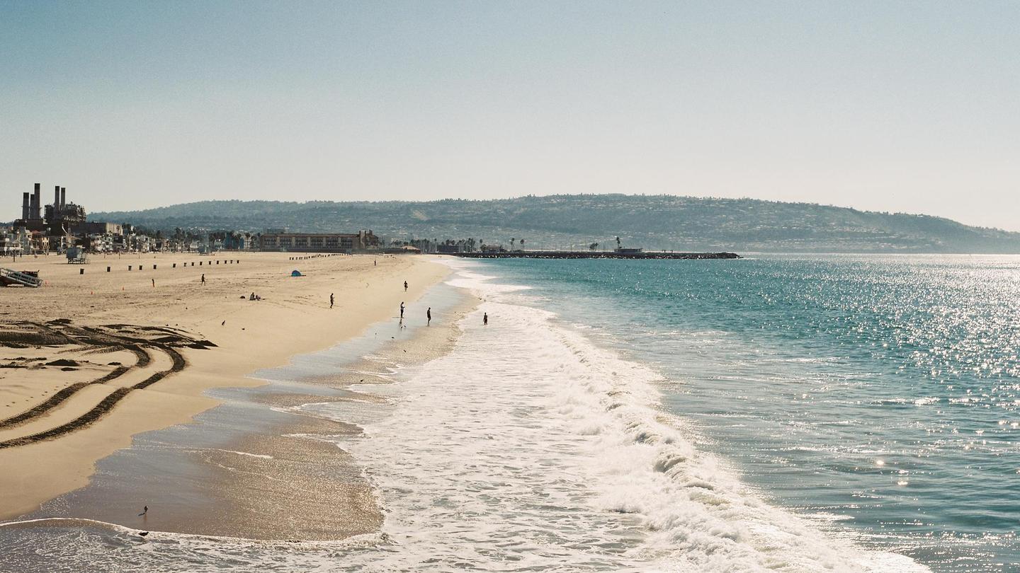 A wide view of Hermosa Beach shows waves breaking on the shore, with buildings and hills in the distance.
