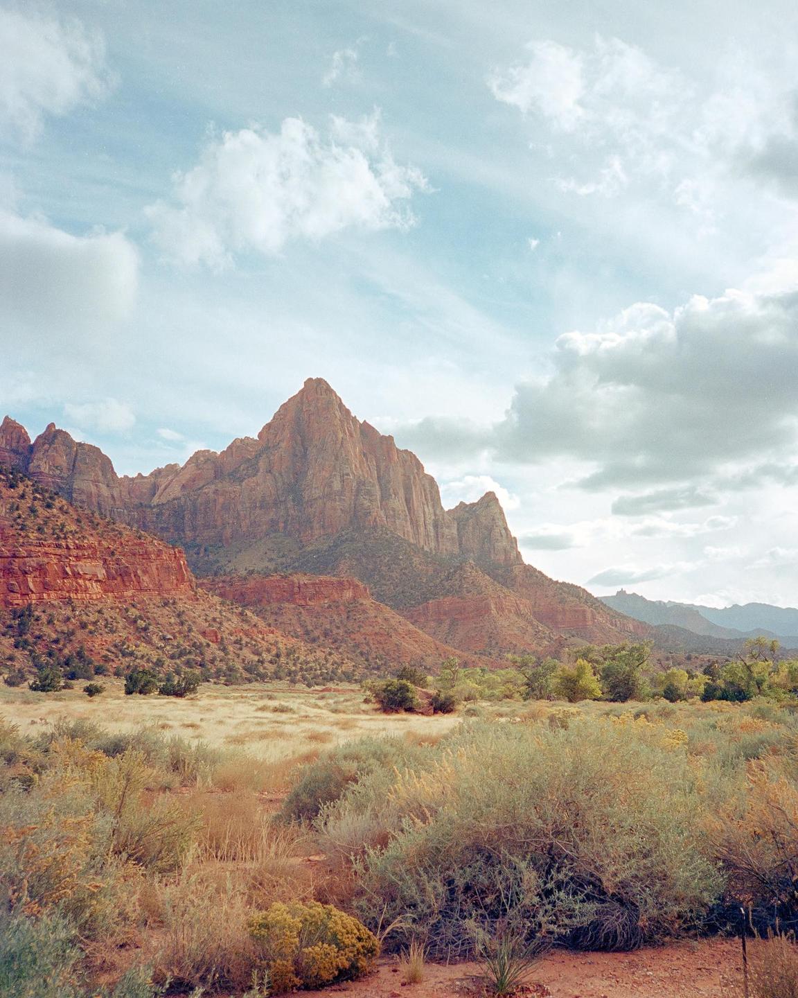 A vast landscape features a prominent red rock mountain under a cloudy sky, with dry brush and green foliage in the foreground.