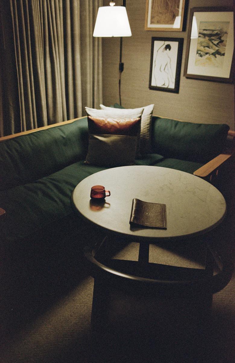 A small red cup and a book are on a round table next to a green sofa with pillows in a dimly lit room.