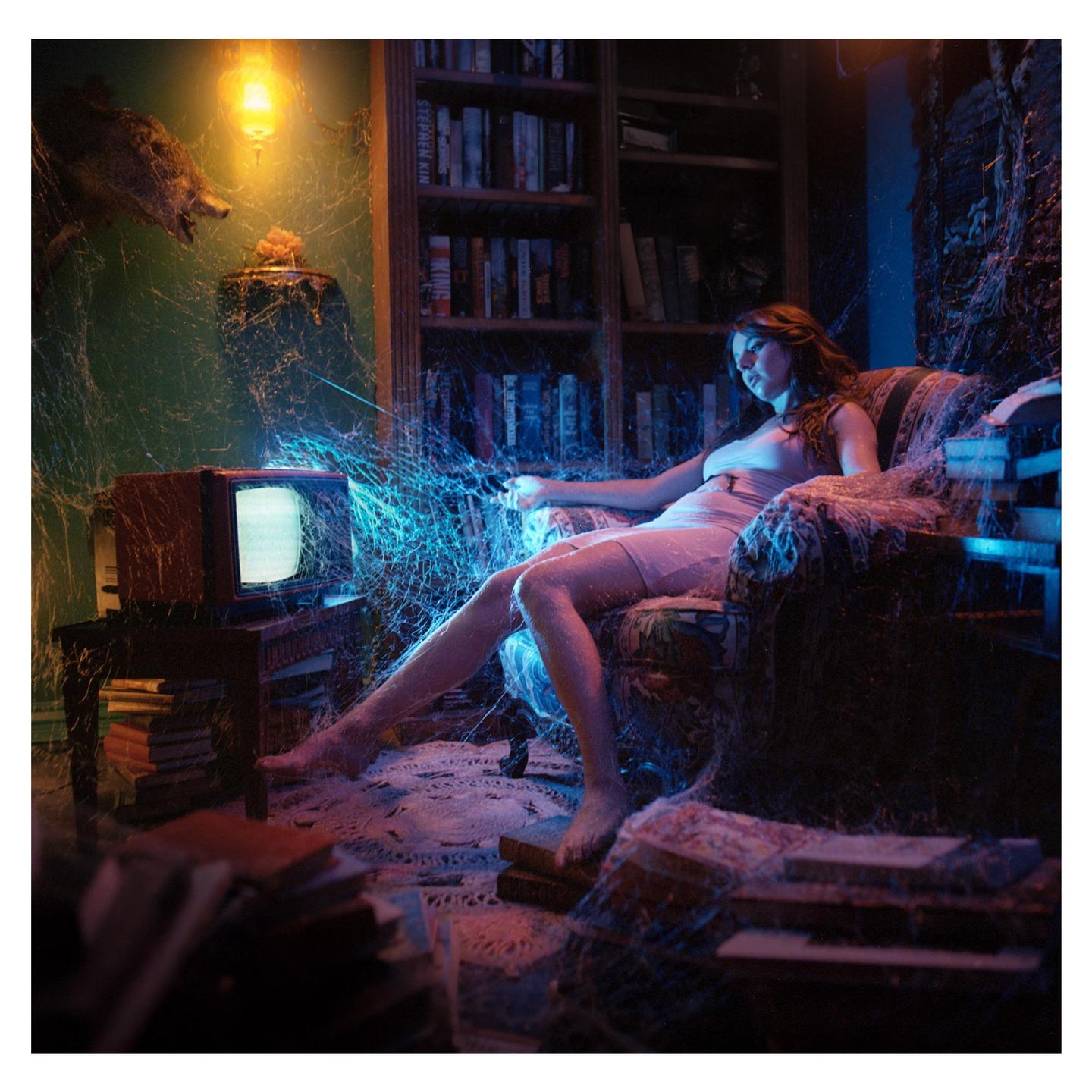 A woman sits in a cobweb-covered armchair, illuminated by a glowing television and a warm light bulb.