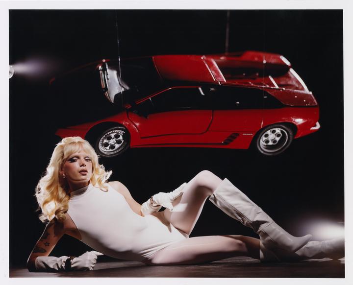 A woman in a white bodysuit and boots reclines on the floor with a red car suspended above her.
