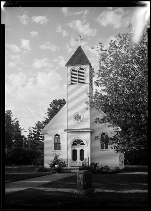 A white church with a steeple and cross stands next to a tree under a cloudy sky.