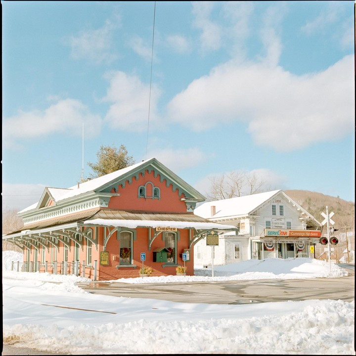 A red brick building with a green trim and a white building with a sign for "Cummings Hardware" are covered in snow under a blue sky with white clouds.