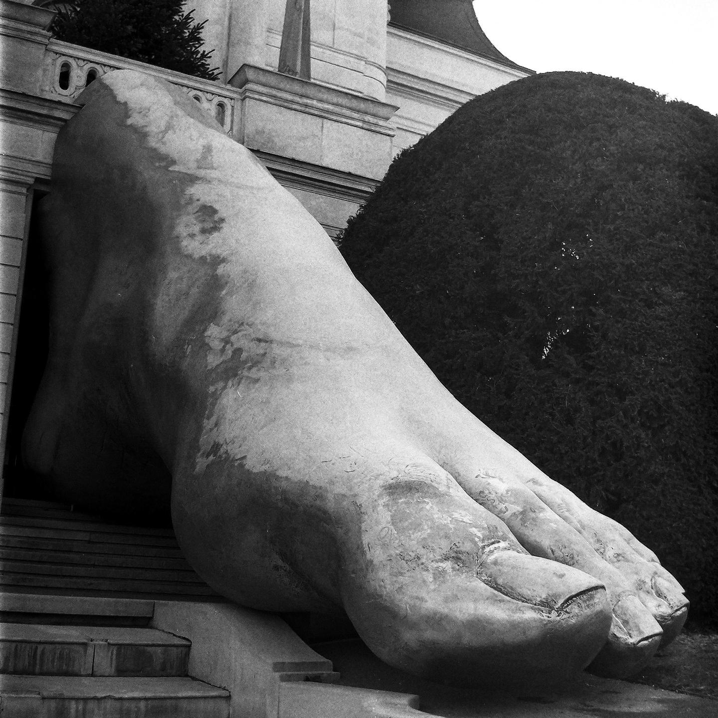 A large sculpture of a foot rests on steps next to a building and a large bush.